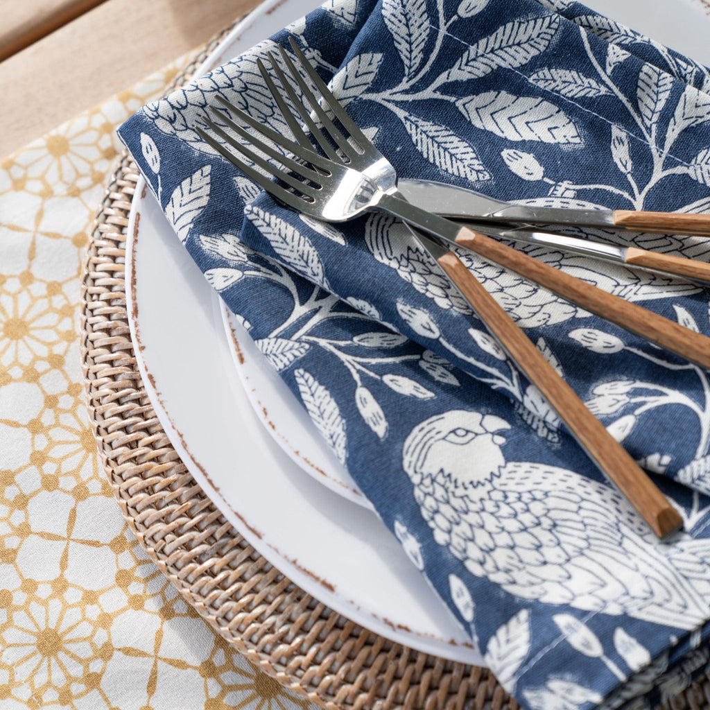 Table setting with a blue and white patterned napkin, silver fork, and wooden spoon on a woven placemat.